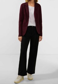 Burgundy blazer med slät yta, enkel knappstängning och två framfickor, i kombination med svarta vida byxor och krämfärgade sneakers.