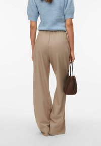 Femme portant un pantalon beige à jambes larges et un haut bleu clair à manches courtes, tenant un petit sac à main marron, vue de dos.