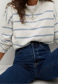 Felpa a righe bianche e azzurre con zip al collo, abbinata a jeans a vita alta di colore blu scuro. Tessuto morbido e vestibilità casual.