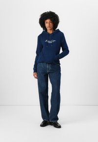 Sweat à capuche bleu marine avec cordons et poche frontale, arborant un logo brodé blanc, associé à un jean en denim foncé à coupe ample.