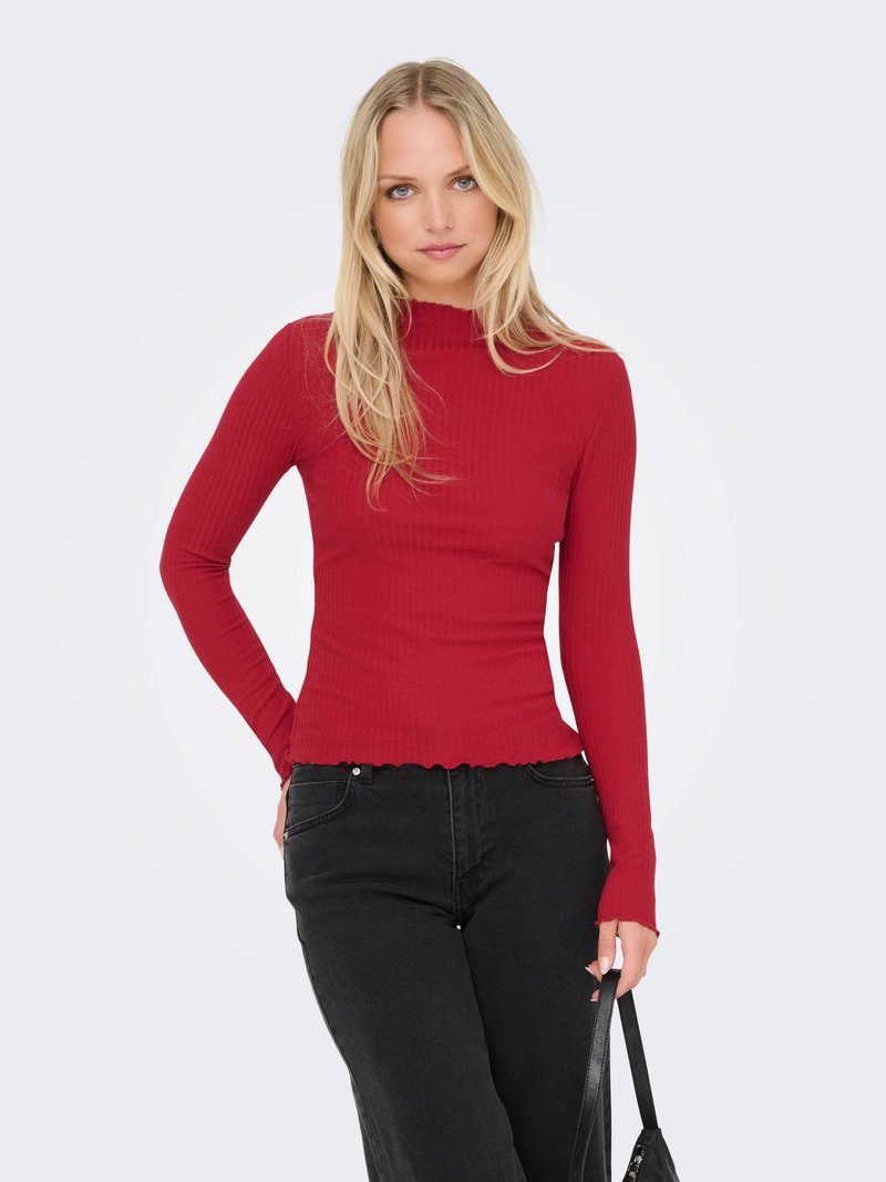 Femme blonde portant un haut moulant rouge à manches longues côtelé et un jean noir, tenant un sac à main noir, debout devant un fond blanc uni.