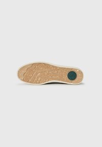 Shoe sole with textured rubber material in beige, featuring a herringbone pattern and a green circular accent near the heel.