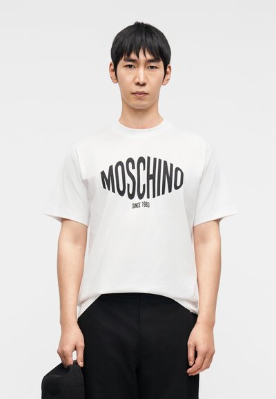 Άσπρο βαμβακερό t-shirt με έντονη μαύρη επιγραφή "MOSCHINO" και λεπτομέρεια "since 1983", σε άνετη γραμμή και με κοντά μανίκια.