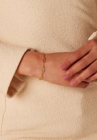 Gouden armband met verlengde schakel, gladde afwerking, gedragen om een pols; aangevuld met zacht beige textuurstof.