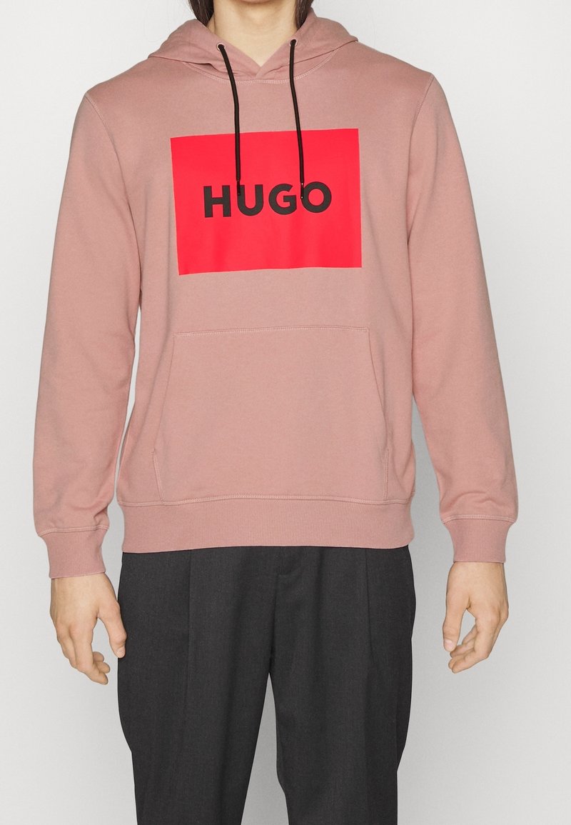 Personne portant un sweat à capuche rose poussiéreux avec un rectangle rouge et le texte noir "HUGO", associé à un pantalon gris foncé, debout sur un fond blanc.