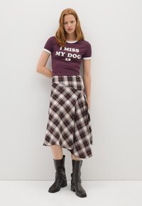 Camisa morada con texto blanco "ECHO DE MENOS A MI PERRO", combinada con una falda envolvente a cuadros en colores rojo, negro y crema. Botas negras hasta la rodilla.