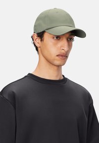 Gorra de béisbol verde con un acabado suave y mate, que presenta paneles cosidos y un visera ligeramente curvada, llevada con un sudadera negra de cuello redondo.