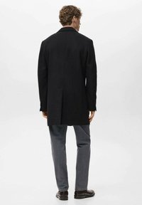 Abrigo de lana negro con abertura trasera y cuello con muesca, combinado con pantalones de mezclilla gris y zapatos de cuero marrón. Diseño limpio y minimalista.