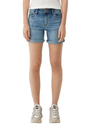 Person iført opkrogede blå denimshorts, hvide sneakers og en hvid top, stående med armene afslappet ned langs siden.