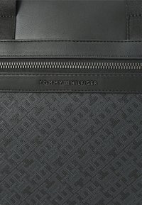 Black textured fabric bag featuring a zippered compartment and embossed "TOMMY HILFIGER" logo. Smooth leather accents are present.