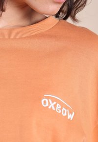 T-shirt en coton orange avec col rond côtelé et logo "Oxbow" brodé en blanc sur le côté gauche. Texture douce et design décontracté.