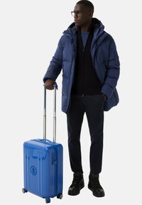 Mężczyzna w okularach i niebieskiej kurtce puchowej stoi, trzymając niebieską walizkę na kółkach z wysuniętym uchwytem na tle białym.