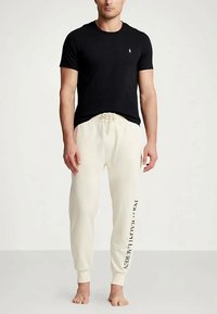 Mann som står barføtt iført en sort t-skjorte med korte ermer og hvite joggebukser med "Polo Ralph Lauren"-tekst på det ene benet.