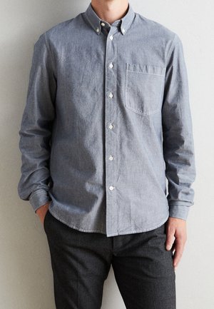 Mężczyzna ubrany w jasnoniebieską koszulę z kieszenią na piersi i ciemne spodnie, stojący z jedną ręką w kieszeni.