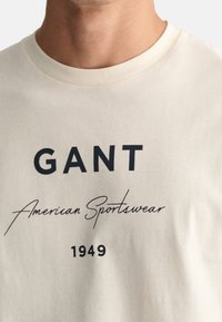 Krémové bavlněné tričko s výrazným černým textem: "GANT American Sportswear" a rokem "1949" v klasickém písmu.
