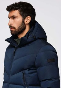 Giacca imbottita blu navy con colletto alto, cerniere e cordini. Presenta una patch con logo sulla manica sinistra e tessuto testurizzato.