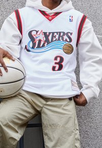 Henkilö valkoisessa Philadelphia 76ers -koripallopaidassa, jonka numero on 3, pitää valkoista koripalloa ja istuu harmaata teksturoitua seinää vasten.