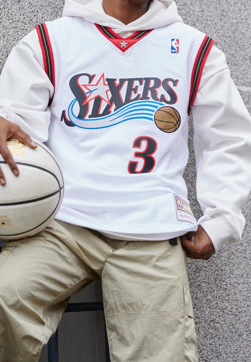 Henkilö valkoisessa Philadelphia 76ers -koripallopaidassa, jonka numero on 3, pitää valkoista koripalloa ja istuu harmaata teksturoitua seinää vasten.