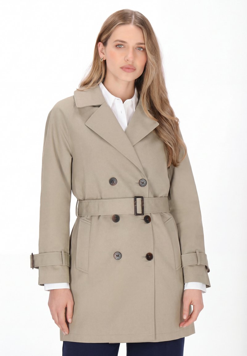 Donna con i capelli lunghi che indossa un trench beige con cintura sopra una camicia bianca, in piedi davanti a uno sfondo bianco semplice.