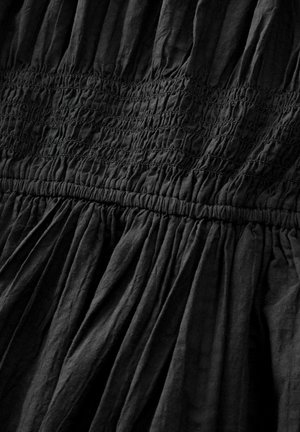 Melnā kleita ar teksturētu, kroku audumu, kas satur savāktus smokošanas elementus jostasvietā un plūstošu svārku. Vienkāršs, minimālisma dizains.