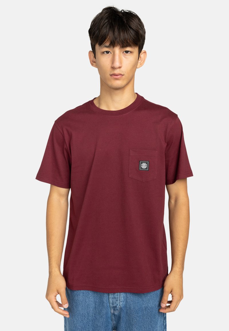 Camiseta de algodón burdeos con cuello redondo, mangas cortas y un bolsillo en el pecho delantero que presenta un logo de etiqueta negra. Textura suave.
