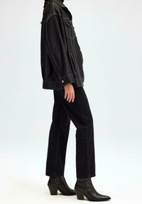 Zwarte faux leren jas met gele stiksels, oversized pasvorm, twee borstzakken; gecombineerd met zwarte rechte jeans en puntige enkellaarsjes.