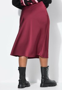 Jupe midi en satin bordeaux avec une silhouette évasée et une texture lisse, associée à des bottes en cuir noir à hauteur de genou ornées de boucles.