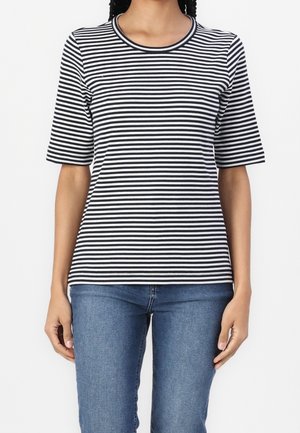 Kvinde iført en sort og hvid horisontalt stribet kortærmet skjorte og blå jeans, der står mod en ensfarvet baggrund.