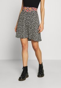 Zwarte bloemenrok met een gemusterde rode en gele tailleband, met een uitlopende snit. Draagt een paar zwarte veterschoenen tot de enkels.