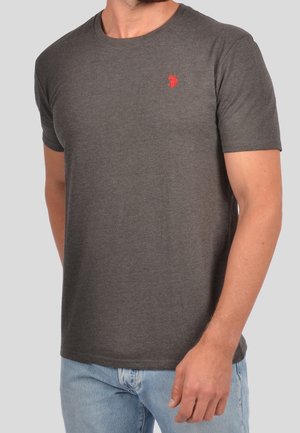 Man draagt een aansluitend donkergrijs T-shirt met korte mouwen en een klein rood logo op de linkerborst en lichtblauwe spijkerbroek.