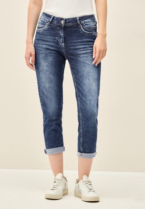 Des jeans en denim de teinte bleue moyenne présentent une coupe slim, des ourlets roulés et des détails de décoloration subtils. Assortis à des sneakers blanches.