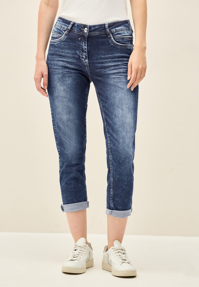 Des jeans en denim de teinte bleue moyenne présentent une coupe slim, des ourlets roulés et des détails de décoloration subtils. Assortis à des sneakers blanches.