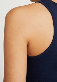 Námořnická modrá bezrukávová tílko vyrobené z jemného bavlněného materiálu, s kulatým výstřihem a hladkou, lehkou texturou. Viditelná ramena a horní část zad.