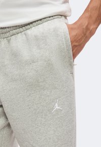 Gros plan d'une personne portant un pantalon de survêtement gris clair avec un logo Jumpman blanc près de la poche, la main partiellement dans la poche.