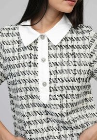 Chemise en tweed texturé noir et blanc avec un col blanc, dotée de trois boutons de fermeture ornés d'accents en argent décoratifs.
