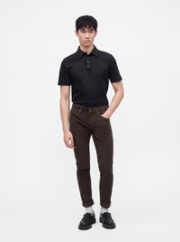 Jeune homme portant un polo noir à manches courtes, un pantalon marron retroussé, des chaussettes blanches et des mocassins noirs, debout avec une main dans la poche.