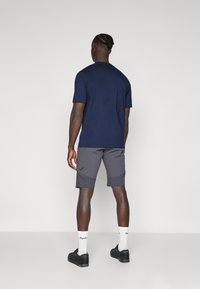 Navy-blaues Kurzarmhemd kombiniert mit anthrazitfarbenen Shorts. Die Shorts haben eine seitliche Tasche und ein Logo-Detail. Schwarze Schuhe und weiße Socken mit Logo.
