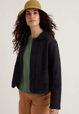 Naine, kellel on lokkis tume juuksed ja kellel on peas beež punutud kübar, must nööpidega jakk, roheline särk ja pruunid püksid, taust on hele ja lihtne.