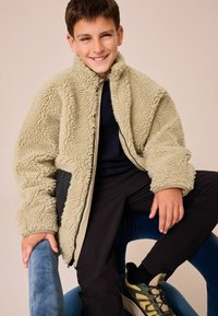 Fleecejacke in hellem Beige mit einer strukturierten, flauschigen Oberfläche; schwarze Akzente entlang der Kanten. Das Modell trägt eine dunkle Hose und grüne Schuhe.