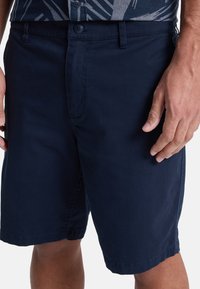 Marineblauwe shorts van lichtgewicht stof. Voorzien van een knoopsluiting, zijzakken en een gladde textuur. De gezoomde randen zijn zichtbaar.