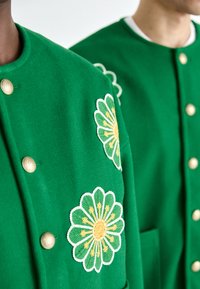 Grønne ulljakker med store blomsterbroderier i gult og hvitt. Gullfargede knapper og en lomme detalj synlig.