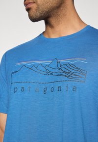 Μπλε βαμβακερό μπλουζάκι με μαύρο γραμμικό σχέδιο βουνών και τη λέξη "παταγονία" σε μαύρο κείμενο κάτω από το γραφικό.