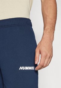 Marinblåa sweatpants har en dragsko i midjan och vit "HUMMEL"-logotyp på benet. Materialet verkar mjukt med en slät textur.
