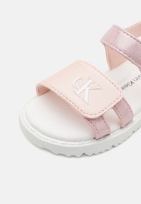 Lys rosa sandal med en myk, glatt tekstur, som har en merket stropp og en hvit, teksturert såle. To justerbare stropper gir en sikker passform.