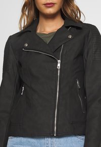 Musta nahkainen moottoripyörätakki, jossa on istuva design, hopeinen vetoketju ja poimuvaljaat harteilla. Sisältää kaksi vetoketjullista taskua.