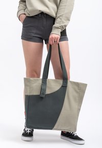 Canvas schoudertas met een grijs en beige kleurblokontwerp, stevige handvaten en een label met een embleem, gehouden door een persoon.