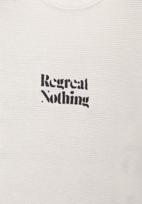 Tissu en tricot blanc avec texte brodé en noir "Regret Nothing" centré sur la zone de la poitrine.