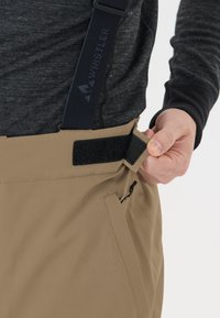Beige Schnee-Hosen mit verstellbaren schwarzen Trägern, ausgestattet mit einem Klettverschluss-Riemen und einer Reißverschluss-Tasche an der Seite. Das Material wirkt glatt und strapazierfähig.