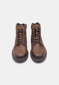 Brązowe skórzane botki za kostkę z okrągłym noskiem, wyposażone w metalowe elementy, oczka, sznurowadła oraz teksturowaną gumową podeszwę dla lepszej przyczepności.