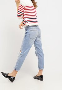 Jeans de denim en un azul claro desgastado con dobladillos enrollados y roturas. Combinados con una blusa de punto a rayas rojas y blancas y zapatos planos negros.